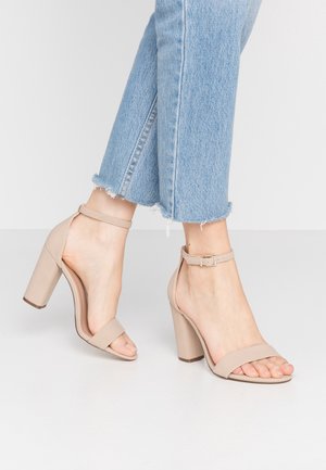 Pieds portant des sandales à talons block beiges avec des brides à la cheville, associés à un jean léger bleu clair à ourlet effiloché, debout sur un sol blanc.