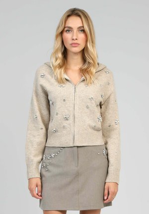 Veste à capuchon beige zippée avec une finition texturée, ornée de motifs de fleurs argentés. Dotée d'un ourlet et de poignets côtelés, portée par-dessus une jupe grise.