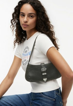Mujer con cabello rizado lleva una camiseta blanca con diseño gráfico y vaqueros azules, sosteniendo un bolso de cuero negro con hebilla plateada.