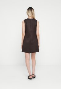 Femme aux cheveux blonds lisses portant une robe noire sans manches et des chaussures noires à talons bas, debout devant un fond blanc uni.