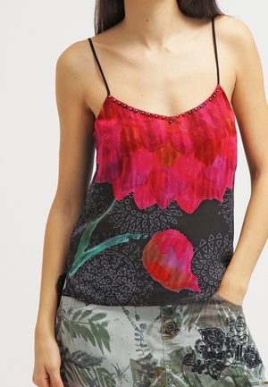 Nainen, jolla on päällään hihaton paita, jossa on ohuet mustat olkaimet, suuri punainen kukkakuosi ja kuviolliset vihreät ja harmaat housut.