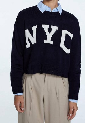 Person wearing a black cropped sweater with white "NYC" letters over a light blue collared shirt and beige pleated pants.