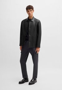 Black leather jacket with a collar and front buttons, paired with striped trousers and black shoes. Smooth texture and tailored fit.