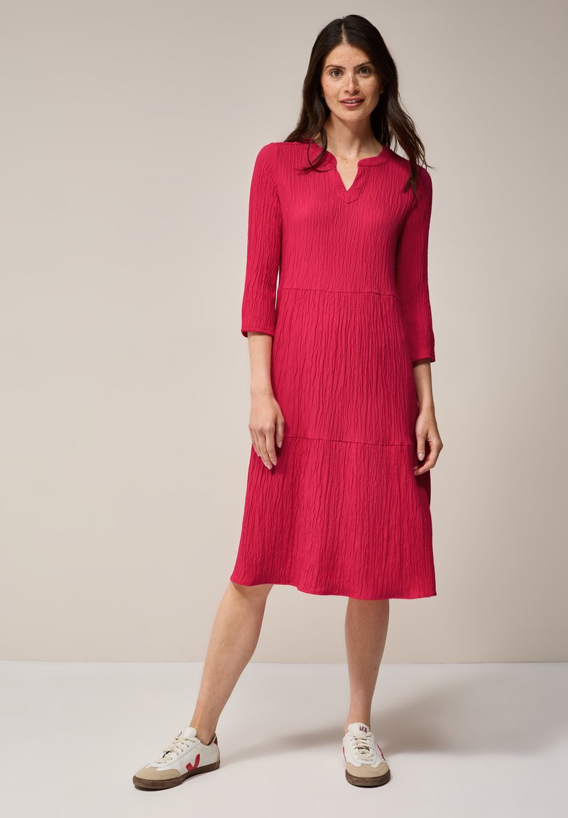 Fuchsia Kleid mit strukturiertem Muster, V-Ausschnitt und drei Viertel-Ärmeln, das knapp unter dem Knie endet; kombiniert mit weißen Sneakers.