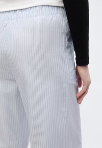 Άτομο που φοράει λευκό παντελόνι με λεπτές κάθετες μαύρες ρίγες και μαύρο μακρυμάνικο τοπ, εμφανίζοντας το δεξί του χέρι να ακουμπάει δίπλα στο σώμα του.