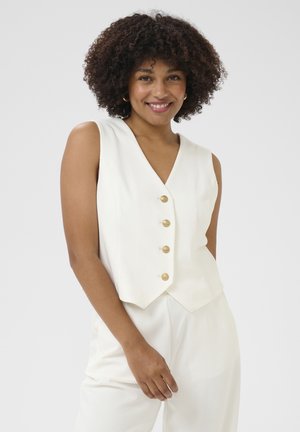 Débardeur blanc sans manches avec un col en V, doté de cinq boutons dorés sur le devant. Texture lisse et coupe ajustée, porté par-dessus un pantalon clair assorti.