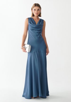 Frau in einem ärmellosen, bodenlangen blauen Satinkleid mit einer silbernen Clutch vor weißem Hintergrund.
