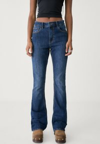 Hoch taillierte, ausgestellte blaue Denim-Jeans mit fünf Taschen, Kontrastnähten und einem leicht verwaschenen Finish. Kombiniert mit braunen Holzclogs.