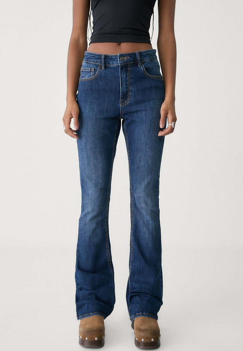 Jean en denim bleu taille haute, évasée, avec cinq poches, des coutures contrastantes et une finition légèrement délavée. Associé à des sabots bruns.