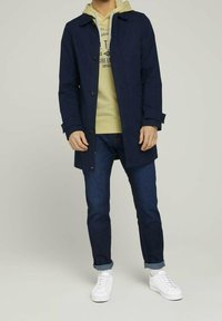Uomo che indossa un cappotto blu navy con bottoni sopra un hoodie beige, jeans scuri arrotolati e sneakers bianche, in piedi contro uno sfondo semplice.