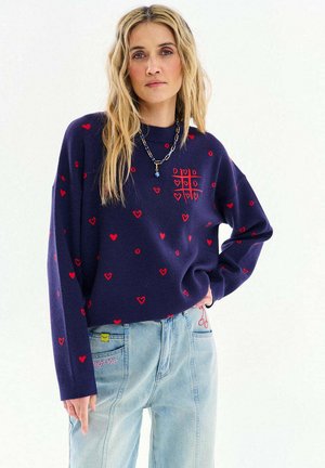 Kobieta o długich blond włosach, ubrana w granatowy sweter z czerwonymi sercami i wzorem kółko i krzyżyk, w połączeniu z jasnoniebieskimi dżinsami z haftowanymi detalami.