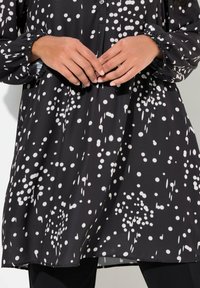Czarna tunika z białym okrągłym wzorem; długie rękawy z marszczonymi mankietami; zapięcie na guziki; lekki, gładki materiał.