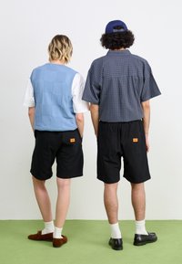 Blau gesteppte Weste über einem weißen Hemd, kombiniert mit schwarzen Shorts. Die Person links trägt braune Slipper; die rechts trägt schwarze Schuhe mit weißen Socken.