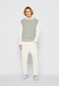 Ljusgrå stickad väst med ett texturerat mönster över en vit skjorta, kombinerat med krämfärgade byxor och ljusblå sneakers. Avslappnad outfit.