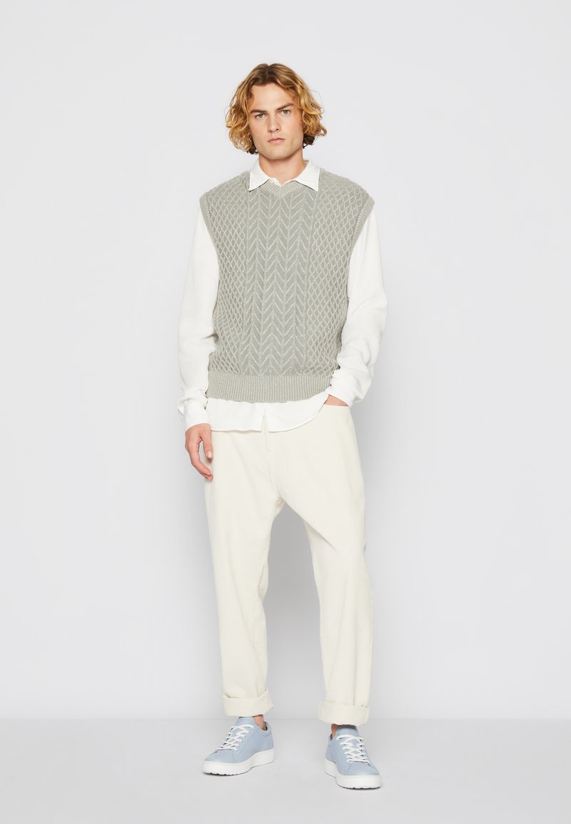 Ljusgrå stickad väst med ett texturerat mönster över en vit skjorta, kombinerat med krämfärgade byxor och ljusblå sneakers. Avslappnad outfit.