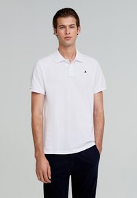 Camisa polo blanca de algodón, con cuello y mangas cortas. Destaca un pequeño logo de calavera negra en el lado izquierdo del pecho. Corte casual.