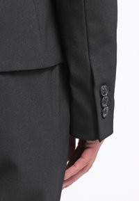 Černá saka s upraveným střihem z hladkého materiálu, s pevným designem a čtyřmi dekorativními knoflíčky na manžetě. Detail rukávu a ruky.