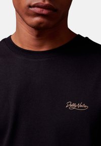 Zwarte katoen t-shirt met een ronde halslijn en een klein crème geborduurd logo op de linkerborst met de tekst "Dolly Noire."