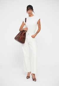 Top blanc à manches courtes, pantalon blanc ample, et grand sac en cuir marron avec des détails en chaîne dorée. Le modèle porte des talons bruns ouverts.