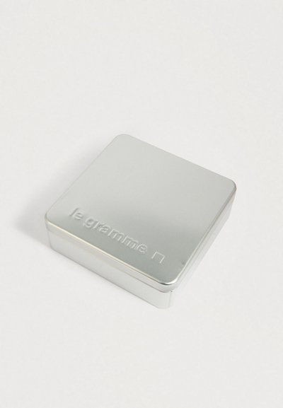 Sudraba metāla kārba ar noapaļotiem stūriem, uz kuras vāka ir izspiestas vārds "le gramme" un neliels kvadrāts, novietota uz baltas virsmas.