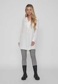 Fehér hosszúujjú blúz gallérral és gomb részletekkel, szürke kockás leggings-szel és térd fölé érő fekete csizmával párosítva.