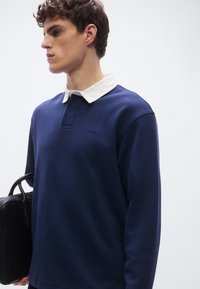 Μακρυμάνικο πόλο σε ναυτικό μπλε με λευκό γιακά, που διαθέτει μερικό πέτο με κουμπιά και διακριτική λεπτομέρεια λογότυπου στο στήθος. Υφασμάτινη υφή.