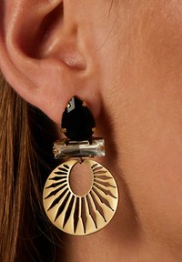 Gouden oorbellen met een zwarte druppelsteen, een heldere rechthoekige steen en een ronde hanger met uitstralende uitsparingen.