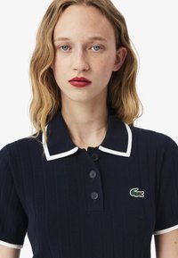 Polo shirt en maille bleu marine avec des bordures blanches et trois boutons ; présente un petit logo de crocodile vert sur la poitrine gauche.