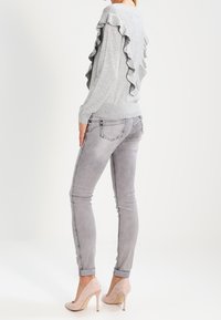 Femme portant un pull à volants gris clair, un jean gris ajusté et des talons hauts rose pâle, debout de profil sur un fond uni.