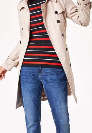 Person iført beige trenchcoat over en sort, hvid og rødstribet sweater med blå jeans mod en ensfarvet hvid baggrund.