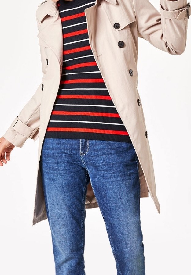 Personne portant un trench-coat beige par-dessus un pull à rayures noires, blanches et rouges avec un jean bleu, sur fond blanc uni.