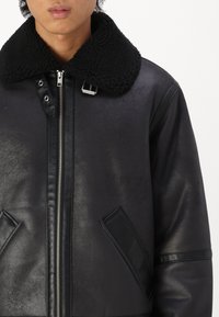 Schwarze Lederjacke mit einem strukturierten schwarzen Schaffellkragen; verfügt über einen vorderen Reißverschluss und schräge Taschen. Glatte, elegante Oberfläche mit matten Akzenten.