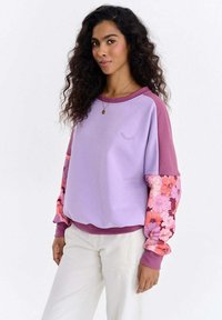 Lilla dressipluus roosade lillemustriliste varrukatega, soonikkoes varrukaotsad, ümar kaelus ja rinnal tikitud silma detail.