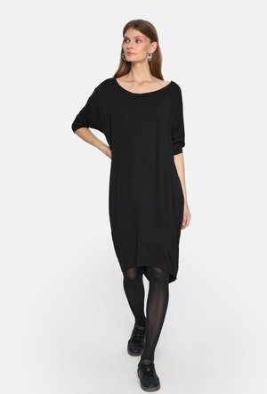 Czarna, oversize sukienka t-shirtowa z szerokim dekoltem i rękawami o długości trzech ćwiartek. Miękka tkanina z delikatnym połyskiem. Noszona z czarnymi rajstopami i butami.