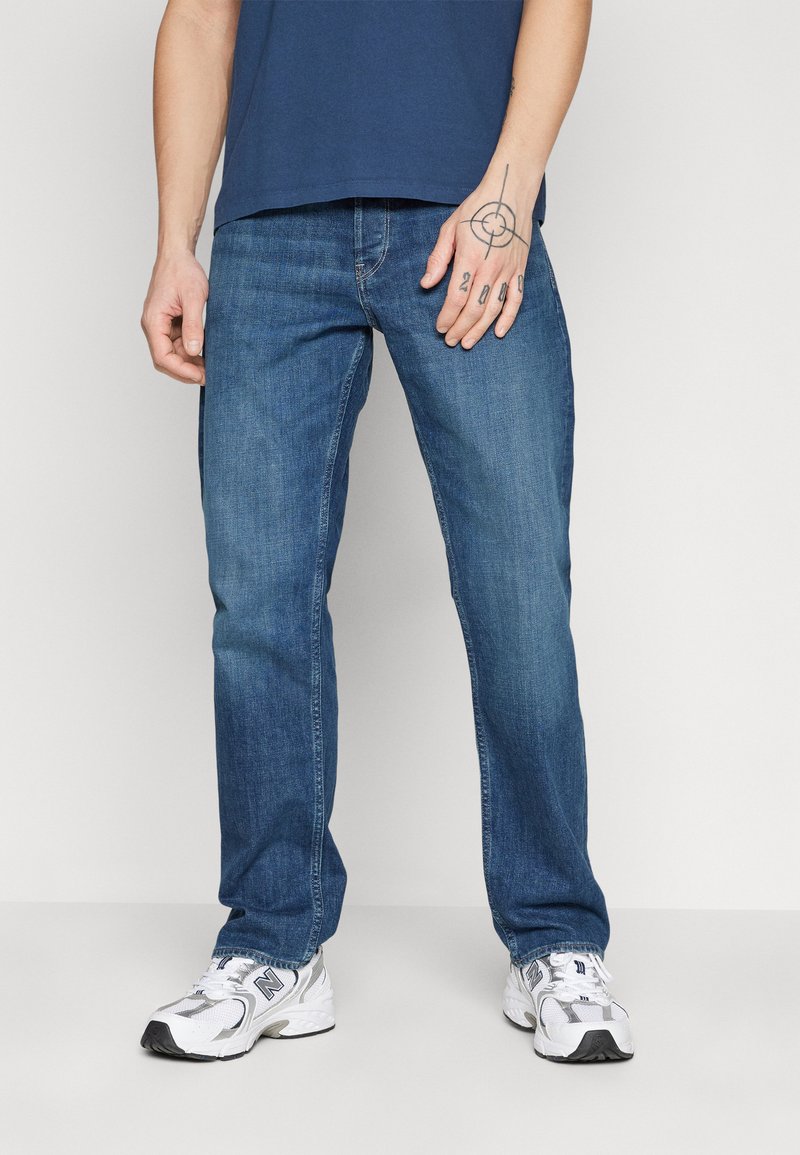 Jean en denim bleu avec une coupe droite, présentant un design à cinq poches et des détails cousus. Accompagnés de baskets blanches.