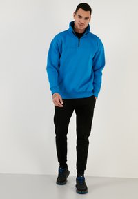 Sudadera azul con cremallera y alto cuello, combinada con pantalones negros ajustados y zapatillas negras con acentos azules. Textura de tela suave.