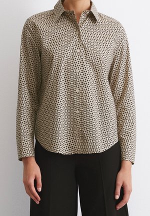 Osoba ubrana w beżową koszulę na guziki z długim rękawem, ozdobioną czarnym geometrycznym wzorem w kształcie koła, oraz czarne spodnie, stojąca na tle jednolitej ściany.