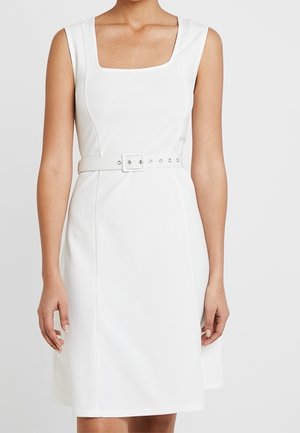 Biała sukienka bez rękawów do kolan z kwadratowym dekoltem i dopasowanym paskiem z klamrą w talii, noszona przez stojącą osobę.