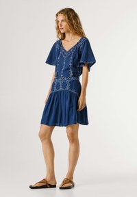 Frau steht und trägt ein blau besticktes kurzes Kleid mit Schmetterlingsärmeln und braunen Sandalen vor einem schlichten, hellen Hintergrund.