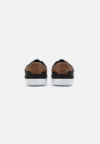 Zapatillas negras con acentos de talón de ante marrón, suelas de goma blanca y parte superior de tela texturizada. La vista trasera muestra una forma elegante y aerodinámica.