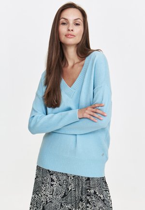 Jasnoniebieski sweter w serek z miękkiej dzianiny, z prążkowanymi mankietami i dołem, w połączeniu z czarno-białą spódnicą w wzory.