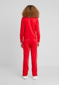 Röd träningsoverall med långa ärmar och matchande byxor. Har vita sidostråk, ribbade muddar och mjuk textur. Bärs med vita sneakers.