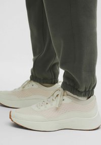 Fehér és világos bézs színű sportcipők texturált hálós kialakítással, gumitalppal és azonos színű fűzős megoldással, sötétzöld szűkített melegítőnadrággal viselve.