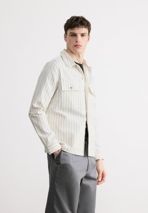 Młody mężczyzna w białej, długiej koszuli w paski zapinanej na guziki i szarych spodniach, stojący z jedną ręką w kieszeni na jednolitym tle.