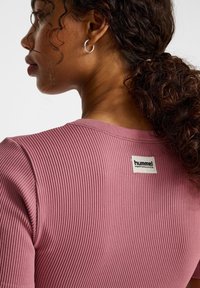 Geribbeld roze shirt met een ronde hals en een label waarop "hummel PERFORMANCE" staat. Krullend haar in een paardenstaart gestyled. Zilveren ringoorbel zichtbaar.
