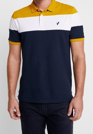 Mann trägt ein kurzärmliges Poloshirt mit horizontalen Streifen in Senfgelb, Weiß und Navy sowie einem kleinen gestickten Vogel auf der Brust.