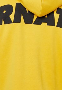 Żółta bluza z kapturem o miękkiej fakturze, z wyraźnym czarnym napisem na klatce piersiowej. Zapinany kaptur z gładkim wykończeniem.