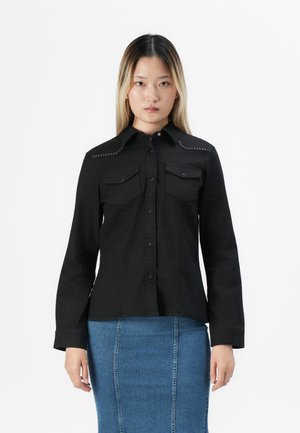 Svart langermet skjorte med to frontlommer, trykknapp-lukking, og dekorative nagler på skuldrene, kombinert med et blått denim-skjørt.