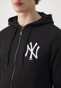 Čierna mikina s kapucňou na zips vyrobená z bavlny, s bielym logom NY na ľavej hrudi, so šnúrkami a textúrovanou tkaninou.