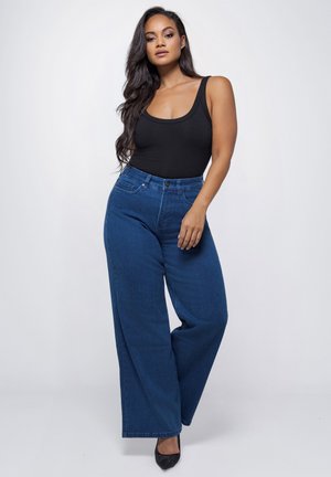 Vrouw met lang donker haar, gekleed in een zwarte mouwloze top, een wijde high-waist blauwe spijkerbroek en zwarte puntige hakken, staand tegen een effen achtergrond.